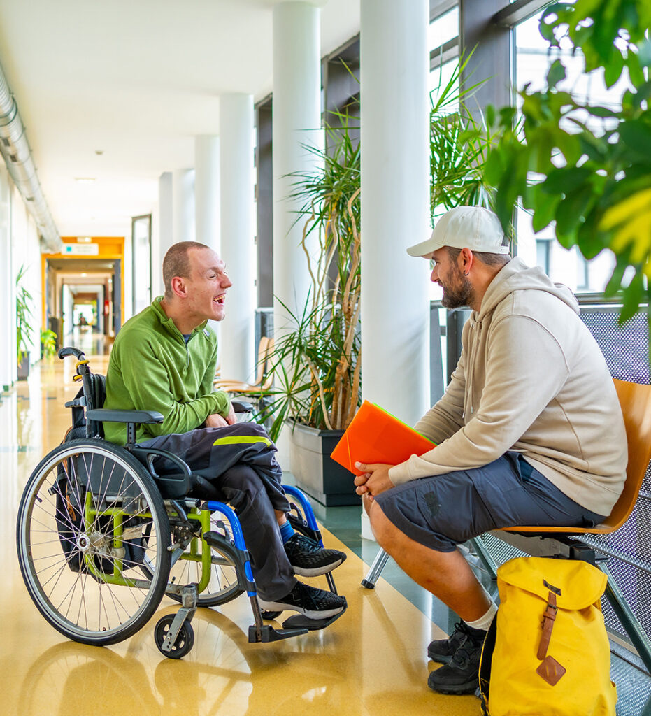 A young man with ALS wearing a green shirt and sitting in a wheelchair laughing with another man in a beige sweatshirt and white hat